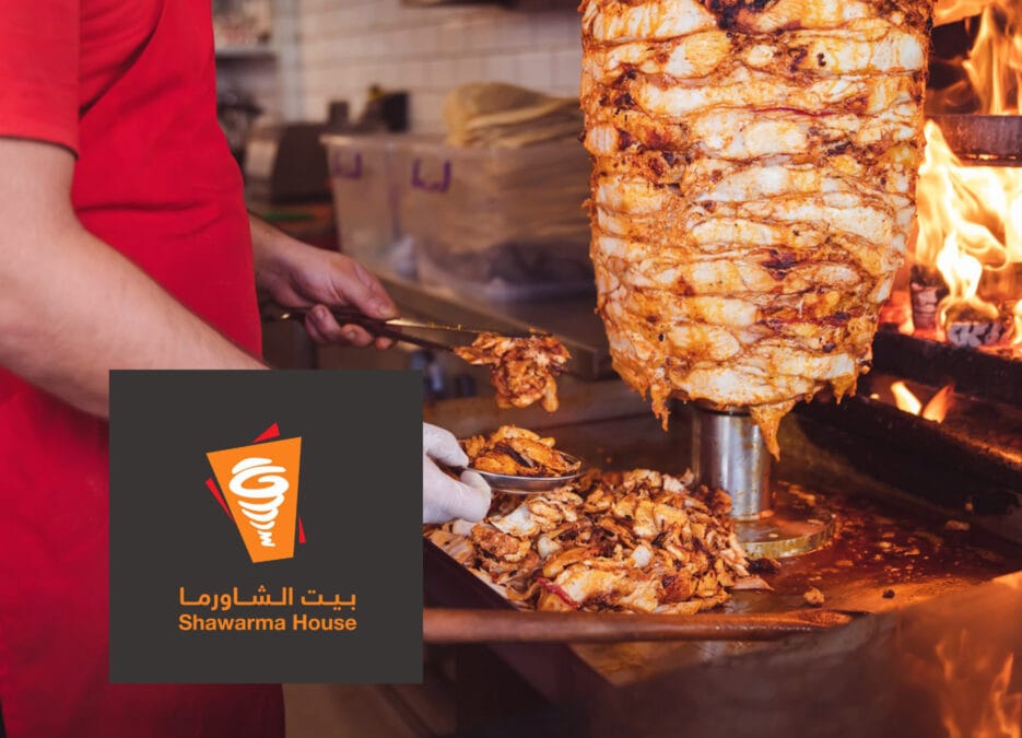 الصورة تضم شخص يقوم بتحضير سيخ من الشاورما، إضافة إلى شعار مطعم بيت الشاورما داخل السعودية
